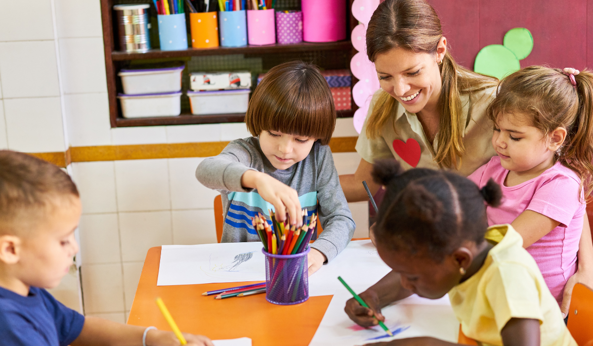 young children drawing at a table with a smiling teacher