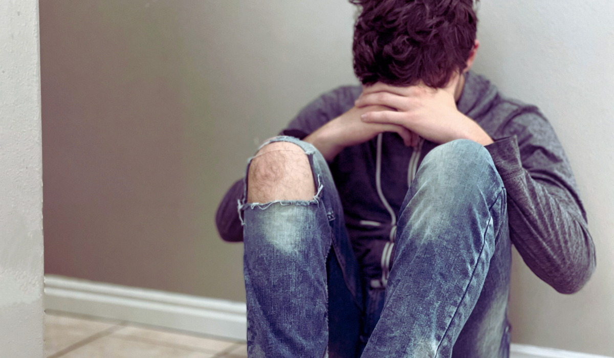 teenage boy in gray hoodie sitting alone in a hallway head in hands depicting sadness, loneliness, stress, emotion