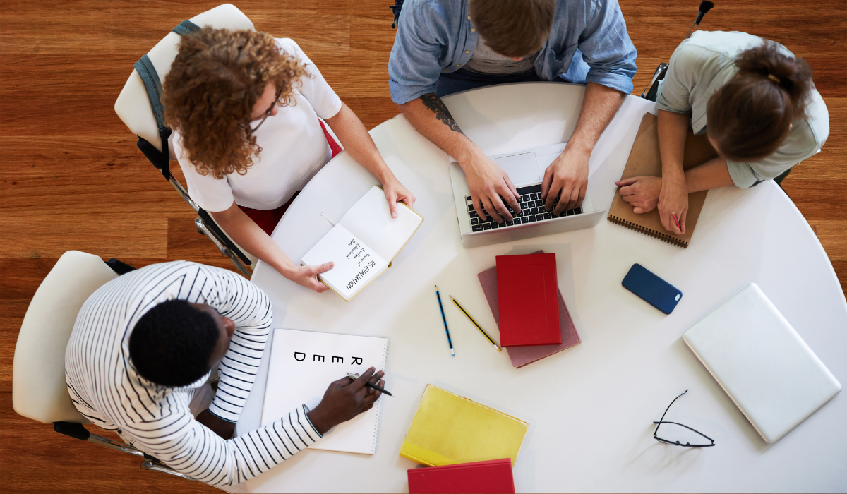 four people sitting around a white table collaborating on a computer with notebooks, pencils
